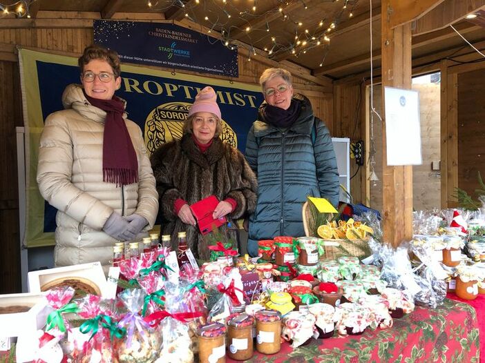Foto vom Stand auf dem Weihnachtsmarkt, im Vordergrund das Angebot an Plätzchen, Marmeladen und anderem Selbstgemachten, im Hintergrund drei Mitglieder des Clubs beim Verkauf